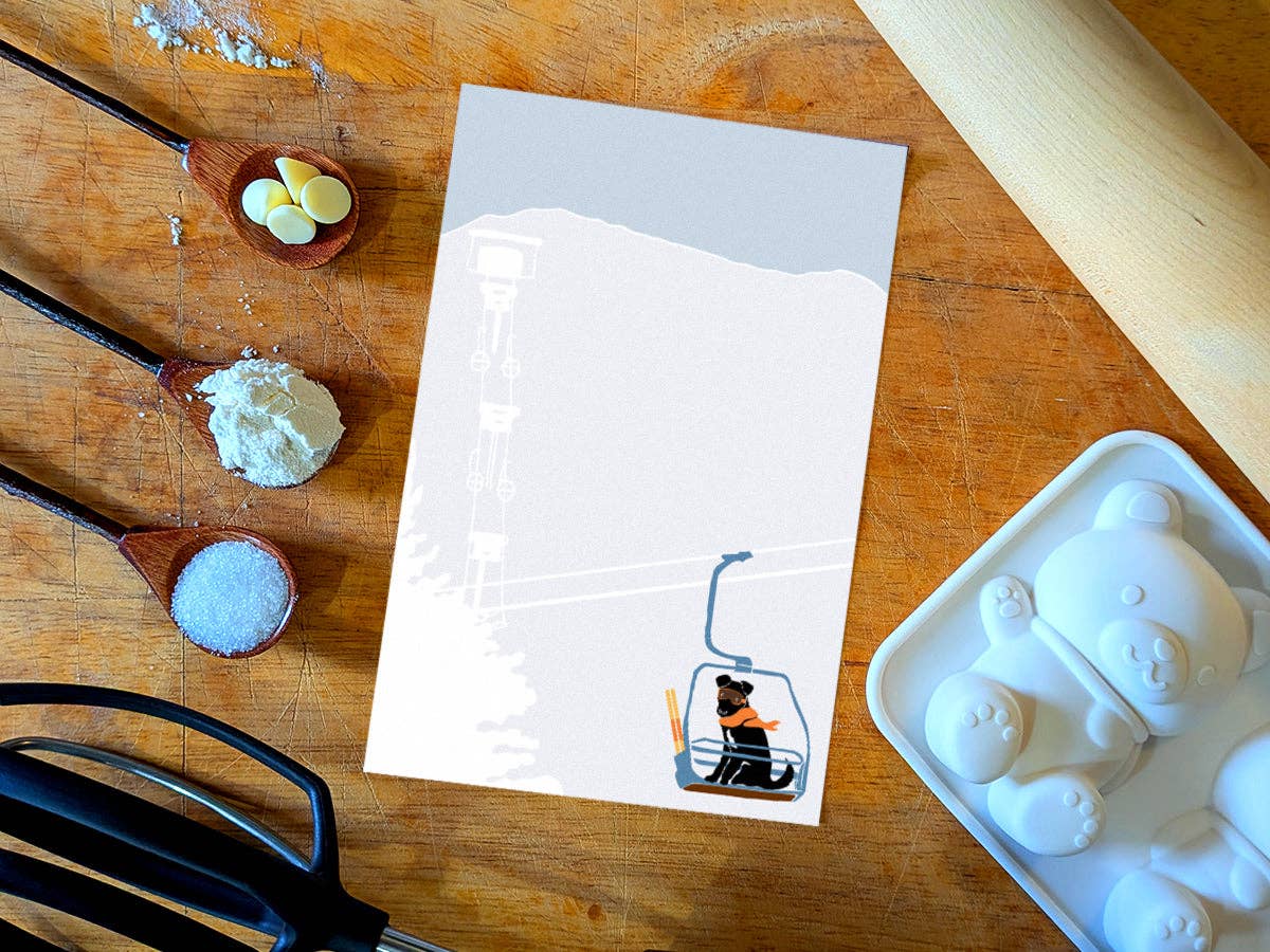 Ski Lift Pup notepad on a wooden counter among baking spoons of white chocolate chips, flour, and sugar next to a bear-shaped silicone mold