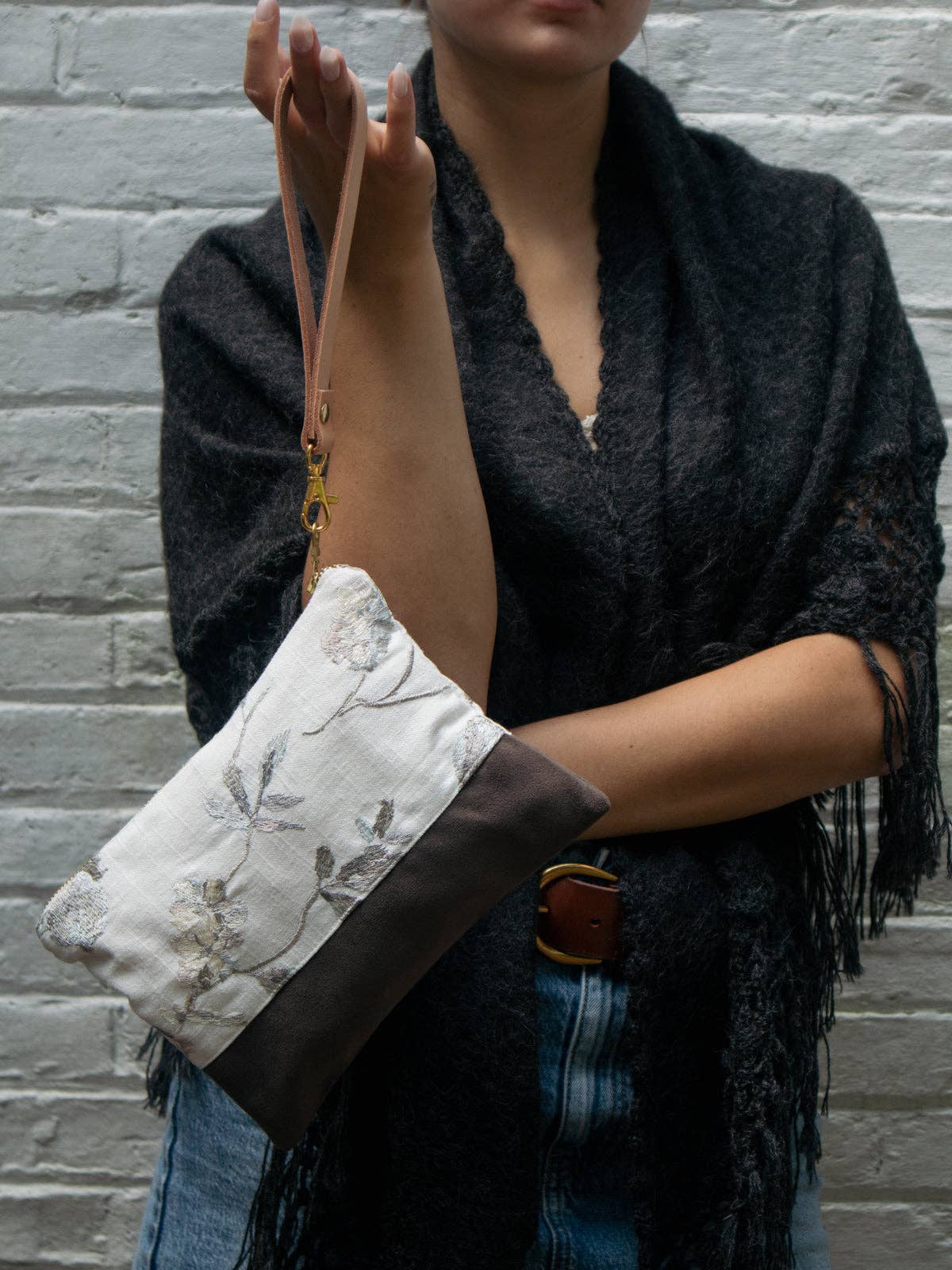 A woman holding a Winona Embroidered Linen wristlet by its tan leather strap, ivory linen with grey floral embroidery over a brown suede base