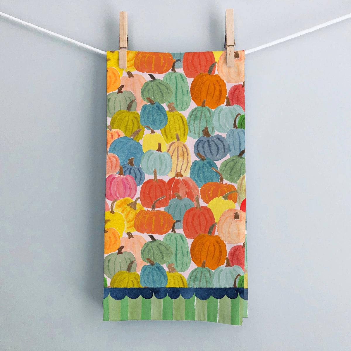 Harvest Pumpkins tea towel hanging from clothespins against a gray wall, with a navy scalloped and green striped hem