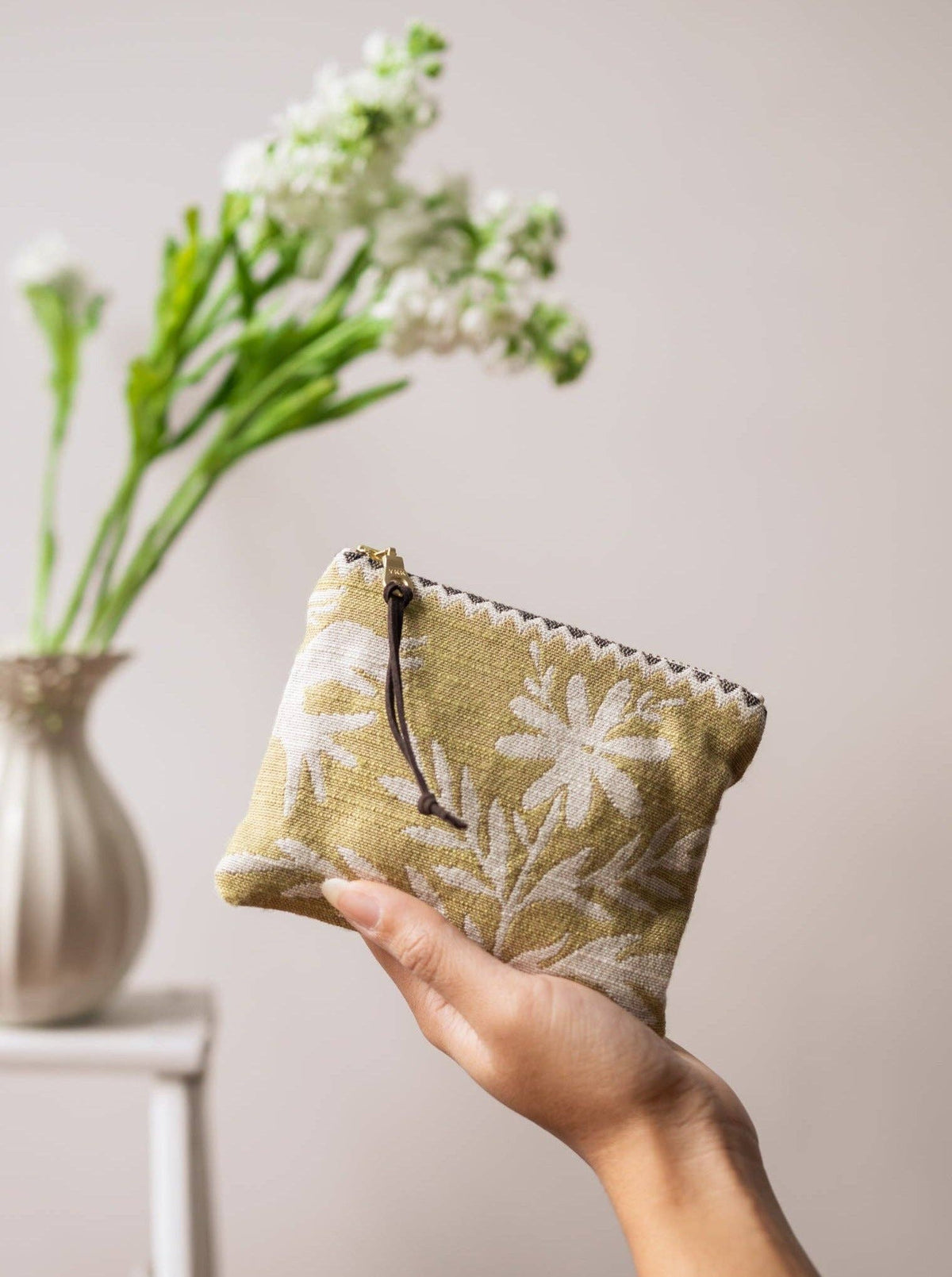 A hand holding a Folk Life mini pouch, olive green with white daisy and fern print, in front of a vase of white flowers