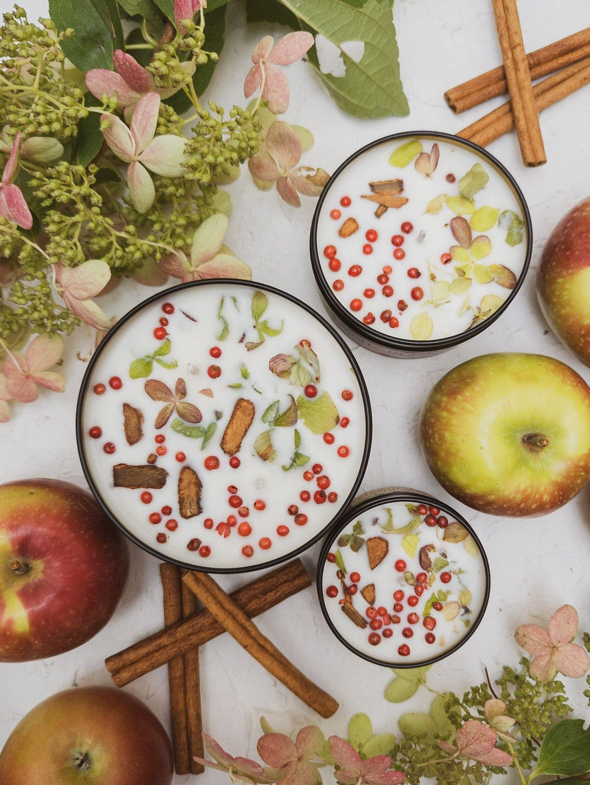 Two HARVEST Apple Spice candle tins of different sizes, wax topped with cinnamon bark and dried hydrangea, surrounded by apples and cinnamon sticks