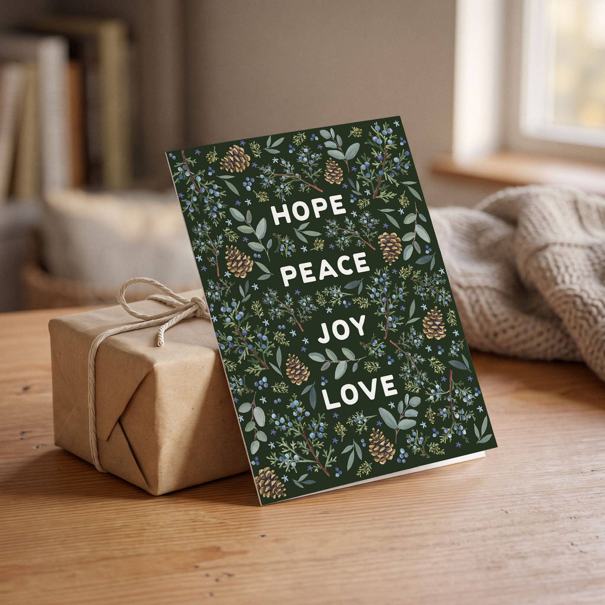Winter Greenery Holiday Card propped against a kraft wrapped gift tied with string on a wood table, juniper berries and pinecones with Hope Peace Joy Love text visible