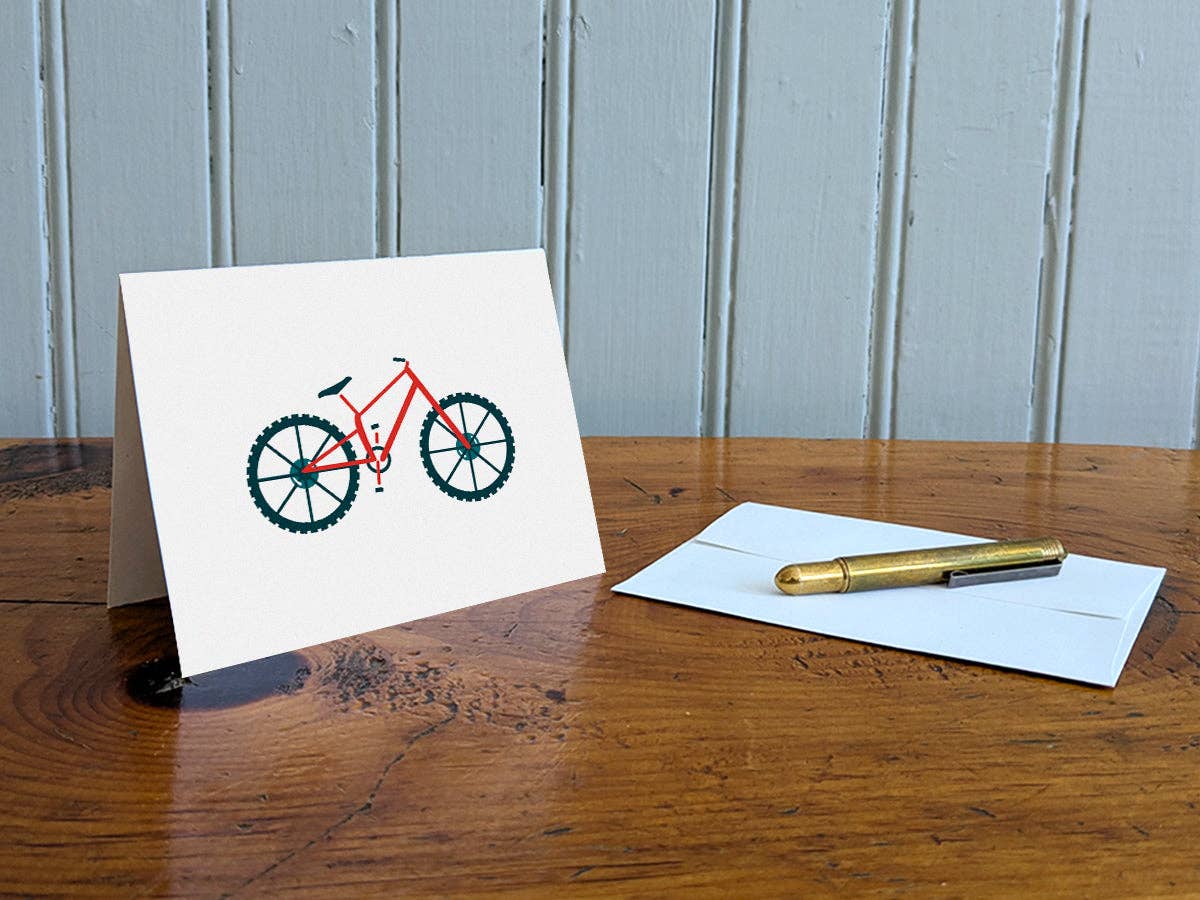 Mountain Bike blank greeting card standing on a wooden table beside a white envelope and a brass pen, with a red mountain bike on the cover
