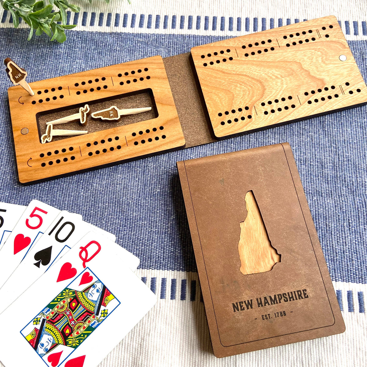 New Hampshire folding travel cribbage board open beside its leather cover, cherry wood board with peg holes and a center slot holding pegs, cover cut out in the shape of New Hampshire and marked New Hampshire Est. 1788, with playing cards