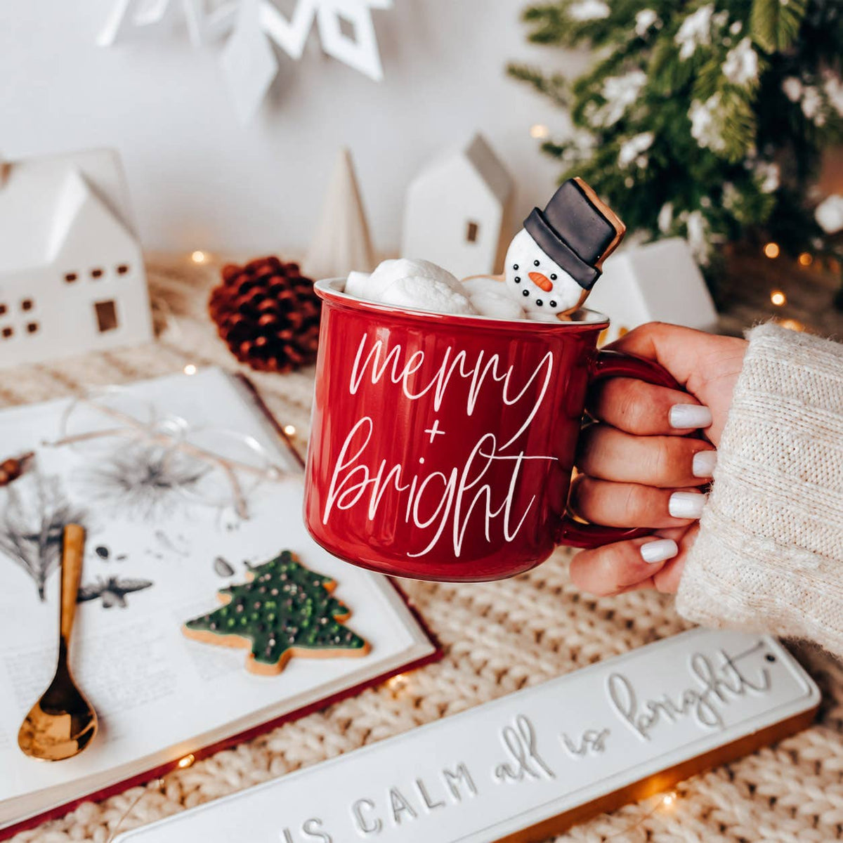 Red campfire-style ceramic mug with merry + bright in white script, held in a hand over marshmallows and a snowman cookie, with white ceramic houses and a pinecone behind