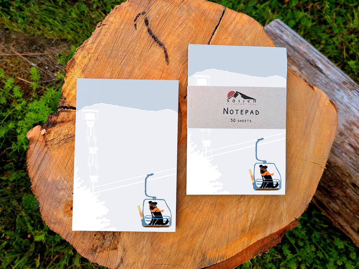 Two Ski Lift Pup notepads on a tree stump, one bare and one wrapped in a kraft paper band reading Soijen Illustrations Notepad, 50 sheets