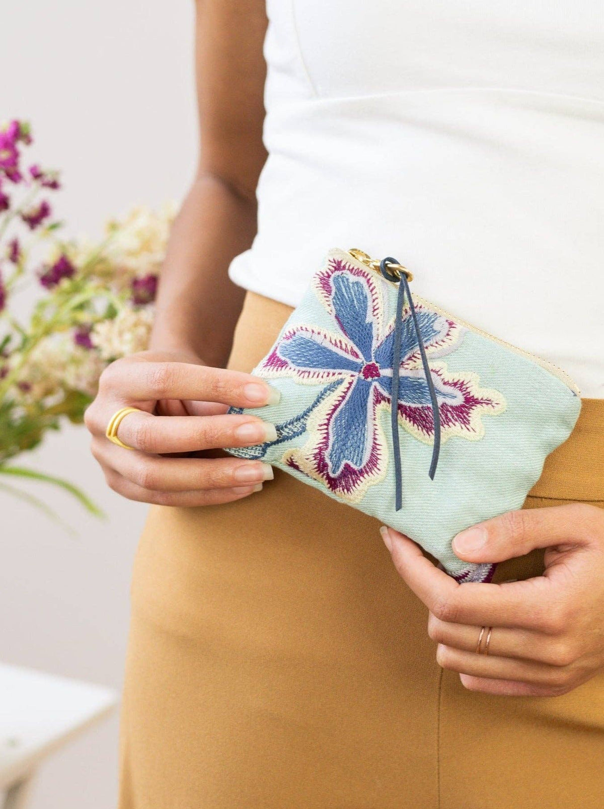 A woman holding an Enchanting Azure embroidered mini pouch against her waist, mint green fabric with magenta and blue floral embroidery
