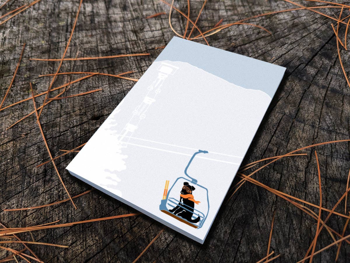 Ski Lift Pup notepad lying on a weathered tree stump with pine needles, showing a black pup in an orange scarf and goggles riding a chairlift
