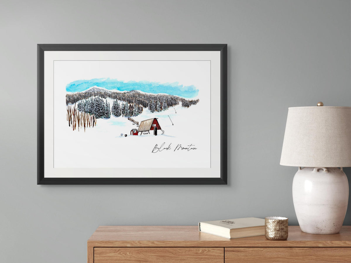 Black Mountain Ski Area print in a black frame, hung above a wooden dresser styled with a lamp, book, and candle