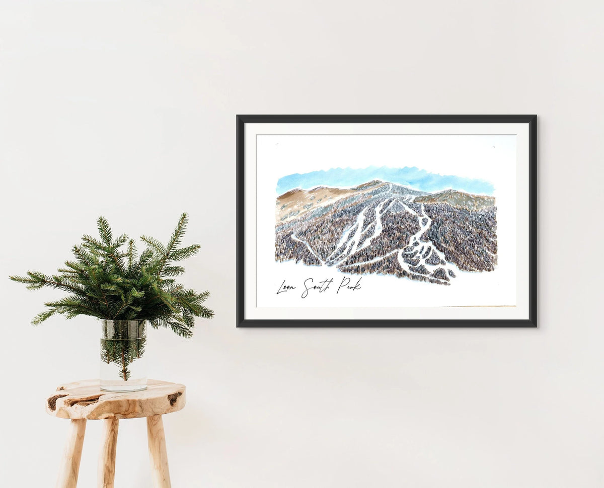 Loon South Peak print in a black frame on a wall, styled with a wooden stool and a vase of pine branches