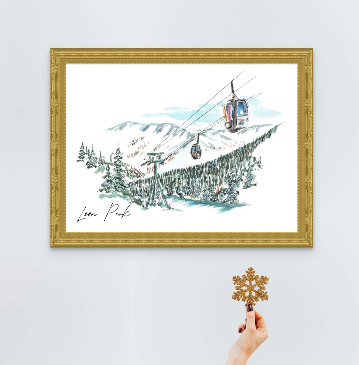 Loon Peak art print in an ornate gold frame, styled with a hand holding a glitter snowflake ornament